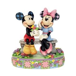 En un banco de ensueño bajo un tapiz de flores multicolor, Mickey regala a Minnie un ramo recién cortado mientras ella sonríe ruborizada, capturando la magia de un momento lleno de ternura gracias al estilo único de Jim Shore. 